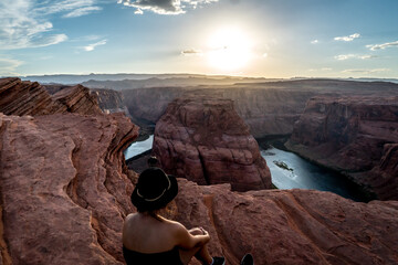 Eine junge Frau sitzt am Rande einer Klippe und schaut dem Sonnenuntergang zu. Horseshoe Bend -...