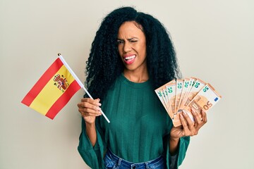 Middle age african american woman holding spain flag and euros banknotes winking looking at the...