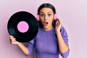 Young hispanic woman using headphones holding vinyl disc afraid and shocked with surprise and...