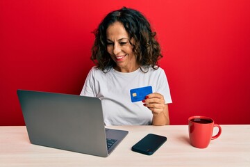 Fototapeta premium Beautiful middle age woman working at the office using laptop and credit card looking positive and happy standing and smiling with a confident smile showing teeth