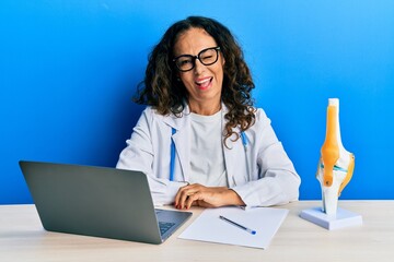 Beautiful middle age woman doctor at orthopedic clinic winking looking at the camera with sexy...