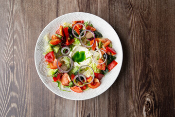 Greek salad with fresh vegetables: tomato, cucumber, red bel pepper, lettuce, onion, olives and cheese. Top view close-up on a white round plate on a wooden background with a copy space. Healthy food.