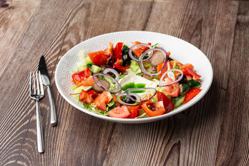 Greek salad with fresh vegetables: tomato, cucumber, red bel pepper, lettuce, onion, olives and cheese. Close-up on a white round plate with cutlery by side on a wooden background. Healthy food.