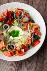A tasty Greek salad with fresh vegetables: tomato, cucumber, red bel pepper, lettuce, onion, olives and cheese. Close-up on a white round plate on a wooden background. Healthy food.