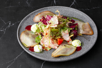 A tasty and sweet salad with pear, tomato, mozzarella cheese and fresh lettuce, dressed with a red and green sauces. Close-up on a grey round plate on a black marble background. Healthy food.