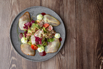 A tasty salad with pear, tomato, mozzarella cheese and fresh lettuce, dressed with a red and green sauces. Close-up top view on a grey round plate on a wooden background with copy space. Healthy food.