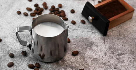 Pitcher with milk surrounded with coffee beans, cup of coffee against concrete background.