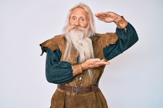 Old senior man with grey hair and long beard wearing viking traditional costume gesturing with hands showing big and large size sign, measure symbol. smiling looking at the camera. measuring concept.