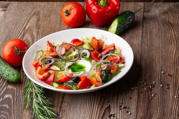 Greek salad with fresh vegetables: tomato, cucumber, red bel pepper, lettuce, onion, olives and cheese. Close-up on a white round plate on a wooden background. Salad menu with ingredients.