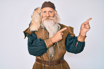 Old senior man with grey hair and long beard wearing viking traditional costume smiling and looking at the camera pointing with two hands and fingers to the side.