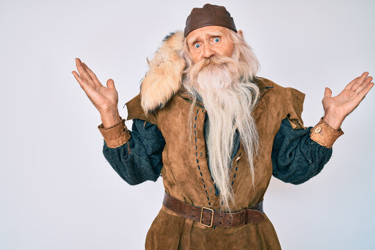 Old senior man with grey hair and long beard wearing viking traditional costume clueless and confused with open arms, no idea and doubtful face.