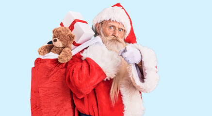 Fototapeta premium Old senior man with grey hair and long beard wearing santa claus costume holding bag with presents pointing with finger to the camera and to you, confident gesture looking serious