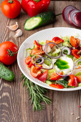 Greek salad with fresh vegetables: tomato, cucumber, red bel pepper, lettuce, onion, olives and cheese. Close-up on a white round plate on a wooden background. Salad menu with ingredients.