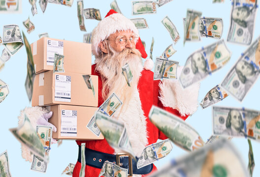Old senior man with grey hair and long beard wearing santa claus costume holding boxes pointing thumb up to the side smiling happy with open mouth