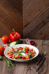 Greek salad with fresh vegetables: tomato, cucumber, red bel pepper, lettuce, onion, olives and cheese. Close-up on a white round plate on a wooden background. Salad menu with ingredients.
