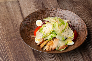 Caesar salad with grilled chicken, tomato, boiled egg, lettuce, croutons and green sauce. Close-up on a brown round plate, isolated on a wooden background with copy space. Healthy food menu.