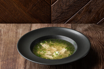 Tasty chicken soup bouillon with noodles, boiled egg, chicken meat, dill and parsley in a black bowl. Isolated close-up on wooden background with copy space. Healthy meal menu. Delicious diet food.