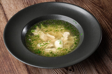 Tasty chicken soup bouillon with noodles, boiled egg, chicken meat, dill and parsley in a black bowl. Isolated close-up on wooden background. Healthy meal menu. Delicious diet food.