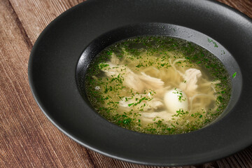 Tasty chicken soup bouillon with noodles, boiled egg, chicken meat, dill and parsley in a black bowl. Isolated close-up on wooden background. Healthy meal menu. Delicious diet food.