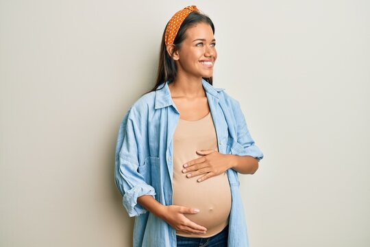 Beautiful hispanic woman expecting a baby, touching pregnant belly smiling looking to the side and staring away thinking.