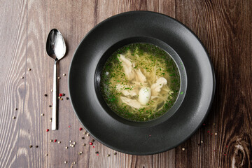 Tasty chicken soup bouillon with noodles, boiled egg, chicken meat, dill and parsley in a black bowl and a spoon by side. Top view close-up on wooden background with copy space. Healthy meal menu.