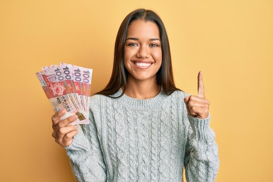 Beautiful hispanic woman holding 100 hong kong dollars banknotes smiling with an idea or question pointing finger with happy face, number one