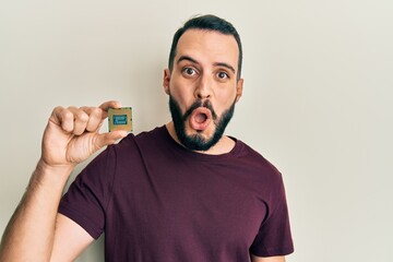 Young man with beard holding cpu computer processor scared and amazed with open mouth for surprise,...