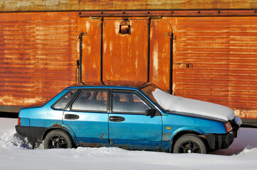 An abandoned car under the snow on the background of an iron wagon