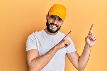 Young man with beard wearing yellow cap smiling and looking at the camera pointing with two hands and fingers to the side.
