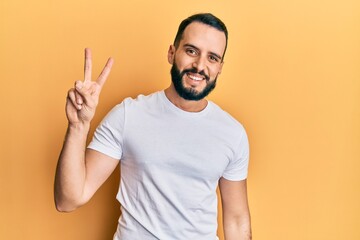Young man with beard wearing casual white t shirt smiling with happy face winking at the camera doing victory sign. number two.