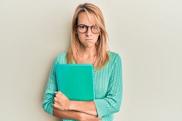 Beautiful blonde woman holding book wearing glasses skeptic and nervous, frowning upset because of problem. negative person.