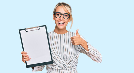 Beautiful blonde business woman holding clipboard with blank space smiling happy and positive, thumb up doing excellent and approval sign