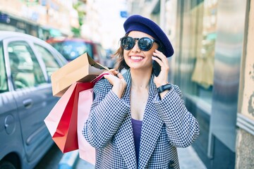 Young beautiful girl with french style holding shopping bags using smartphone at street of city