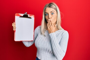 Beautiful blonde woman holding clipboard with blank space covering mouth with hand, shocked and...