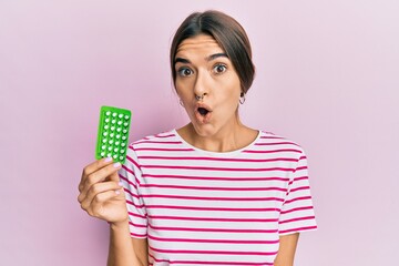 Young hispanic woman holding birth control pills scared and amazed with open mouth for surprise,...