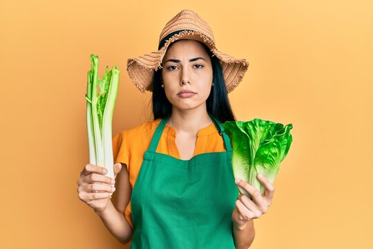 Beautiful young woman wearing gardener apron holding vegetables relaxed with serious expression on face. simple and natural looking at the camera.
