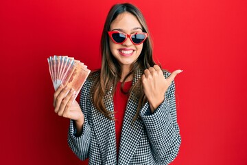 Young brunette woman holding 100 norwegian krone banknotes pointing thumb up to the side smiling...