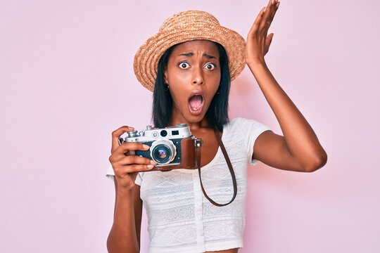 Young african american woman wearing summer hat holding vintage camera surprised with hand on head for mistake, remember error. forgot, bad memory concept.