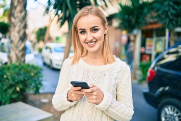 Young blonde girl smiling happy using smartphone at the city.