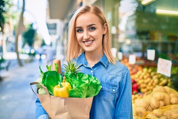 Young blonde girl smiling happy holding groceries paper bag standing at the supermarket