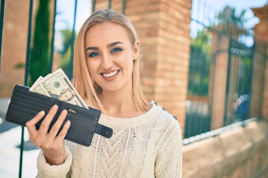 Young blonde girl smiling happy holding wallet with dollars standing at the city.