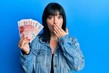 Young hispanic woman holding icelandic krona banknotes covering mouth with hand, shocked and afraid...