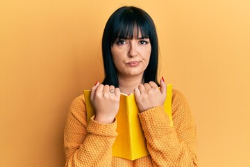 Young hispanic woman holding book skeptic and nervous, frowning upset because of problem. negative...