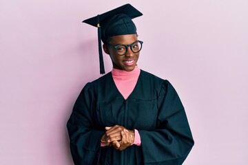 Young african american girl wearing graduation cap and ceremony robe with hands together and crossed fingers smiling relaxed and cheerful. success and optimistic