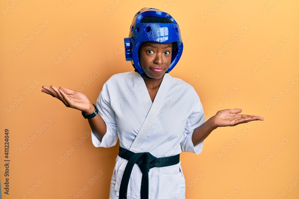 Poster Young african american girl wearing taekwondo kimono and protection helmet clueless and confused with open arms, no idea and doubtful face.