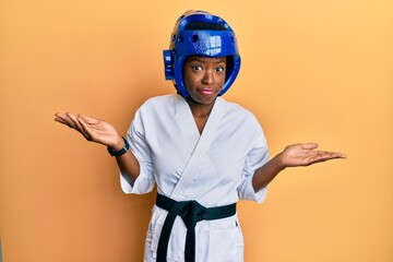 Young african american girl wearing taekwondo kimono and protection helmet clueless and confused with open arms, no idea and doubtful face.