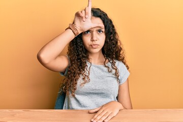 Teenager hispanic girl wearing casual clothes sitting on the table making fun of people with fingers on forehead doing loser gesture mocking and insulting.