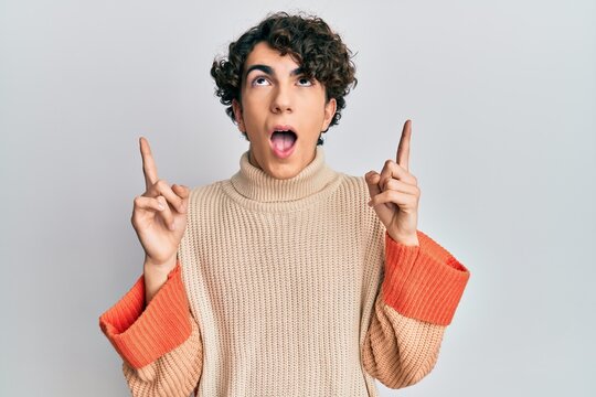 Hispanic young man wearing casual winter sweater amazed and surprised looking up and pointing with fingers and raised arms.