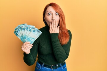 Beautiful redhead woman holding 100 brazilian real banknotes covering mouth with hand, shocked and...