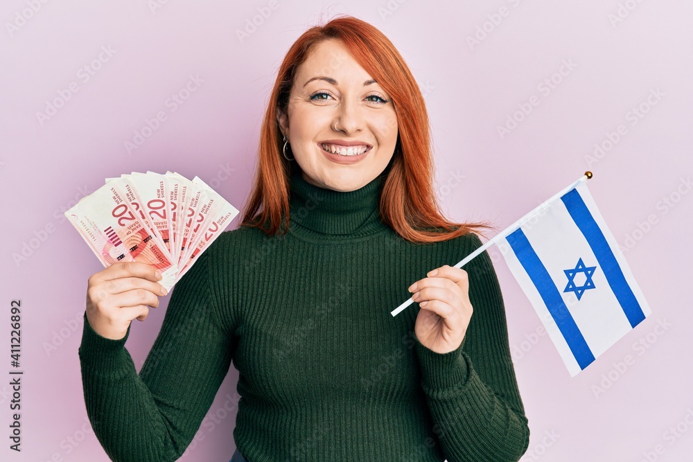 Poster beautiful redhead woman holding 20 shekels banknotes and israel flag smiling with a happy and cool s